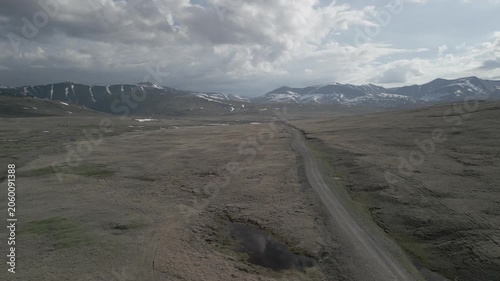 Low altitude drone flight along mountain dirt road with puddles reflecting snowy peaks in spring valley. Travel journey. RAW flat profile uncorrected video footage.