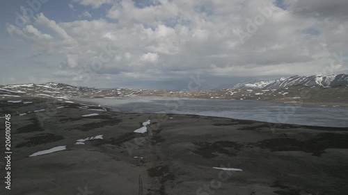 Aerial shot of dirt road leading to frozen alpine lake in high mountains during spring thaw. Vast landscape with snowy peaks. RAW flat profile uncorrected video footage.