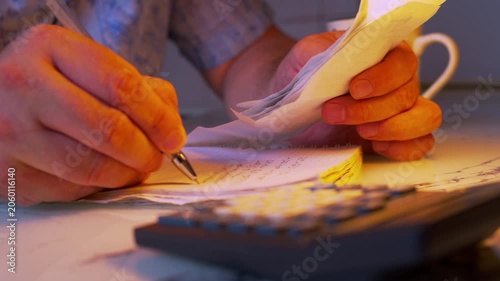Cinematic close-up shot of writing down expense numbers from bills into a ledger book. Dark room, warm lamp light. Visualizes tax preparation, financial audit, managing personal debt and money savings