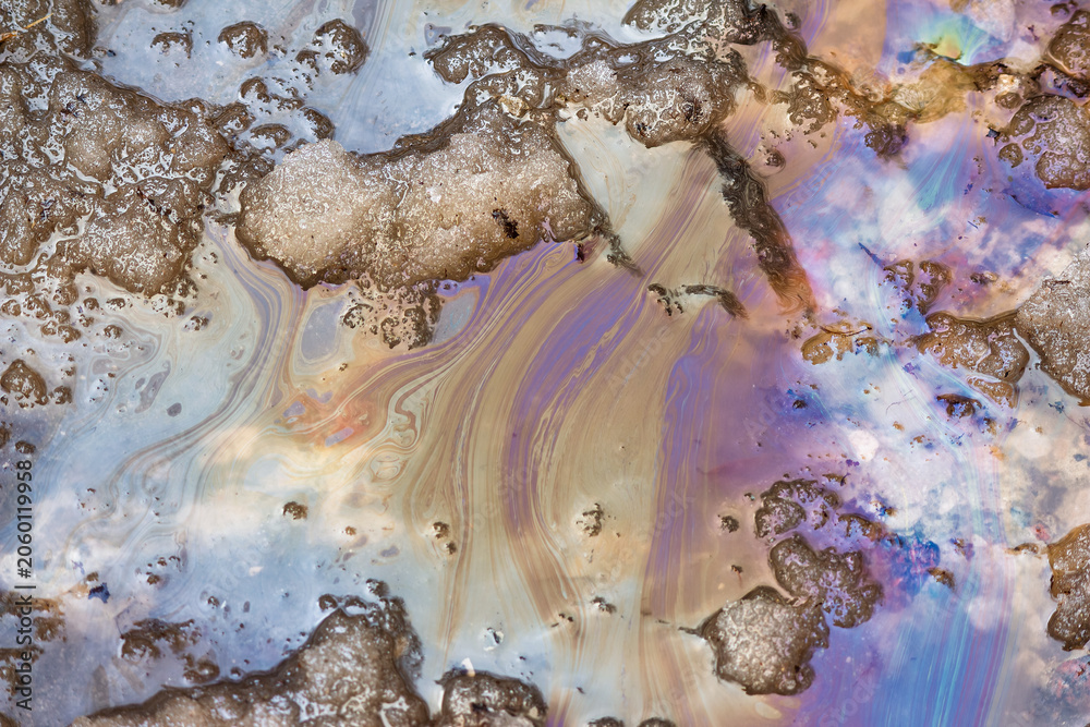 Fototapeta premium Iridescent oil slick patterns swirling on melting snow during spring thaw, forming colorful toxic gasoline and petroleum residues in a dirty slush puddle on the asphalt road surface