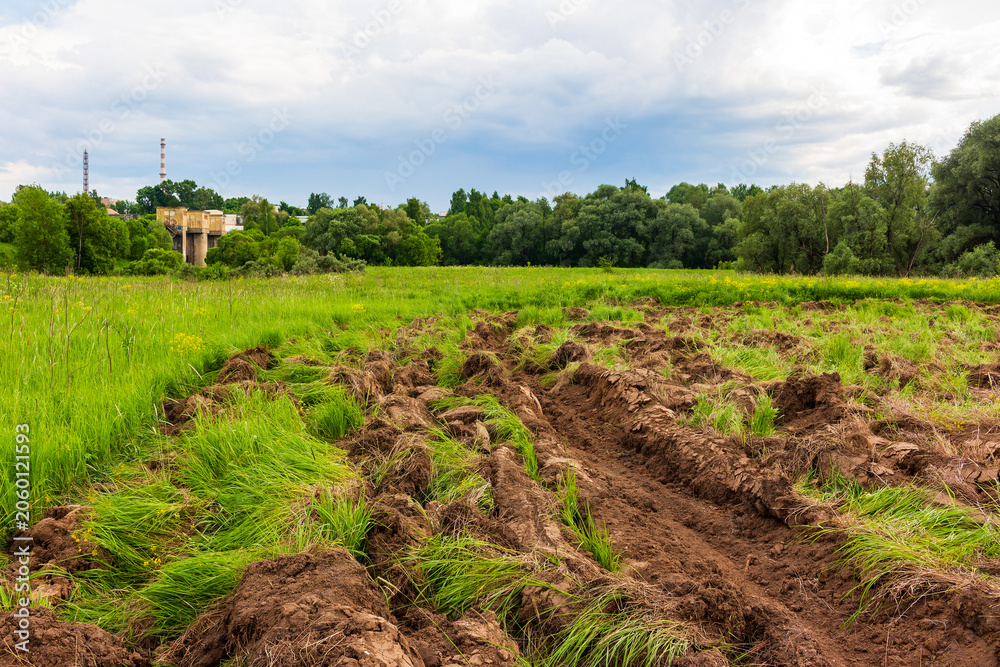 Obraz premium Freshly plowed brown soil furrows cutting through a vibrant green overgrown meadow, showcasing the raw process of reclaiming wild land for future agricultural planting under cloudy skies