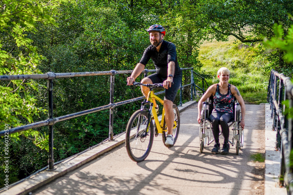 Obraz premium Man cycling and woman in wheelchair enjoying a sunny day on a bridge.