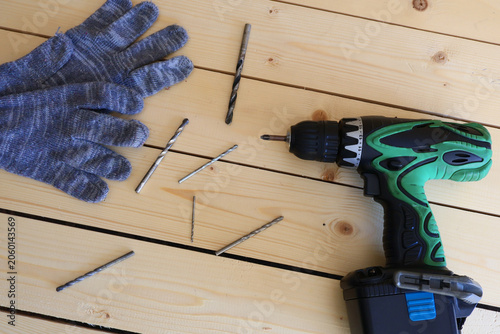 Green cordless drill lies next to scattered drill bits and a pair of blue work gloves on light pine wood. Tools are neatly arranged, signaling preparation for a home improvement or woodworking task. C