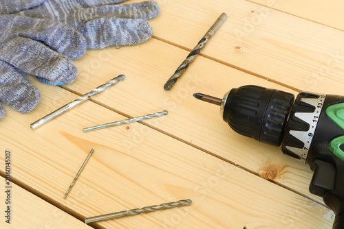 Green cordless drill lies next to scattered drill bits and a pair of blue work gloves on light pine wood. Tools are neatly arranged, signaling preparation for a home improvement or woodworking task. C