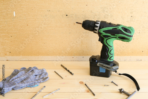 Green and black cordless drill stands ready on light wood surface, surrounded by drill bits, screws, and protective gloves. Clean, organized setup conveys efficiency, preparation, and safety for home 