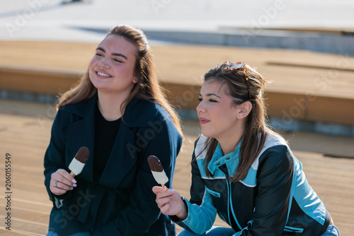 Two young women enjoy ice cream while sitting outdoors on bench