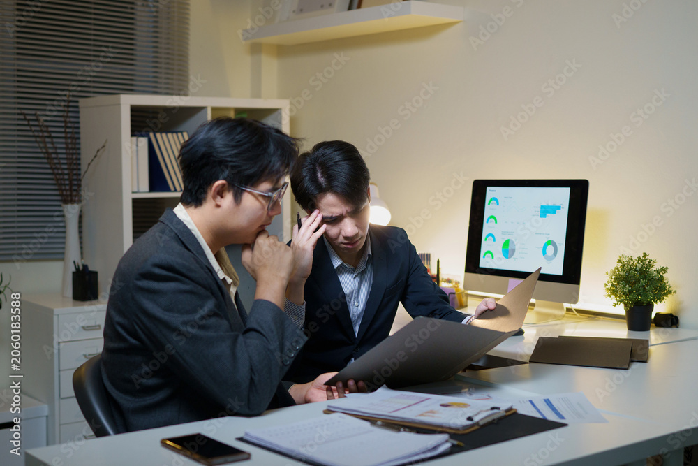 Fototapeta premium Two Asian Business Professionals Reviewing Documents and Data Charts Late at Night in Modern Office
