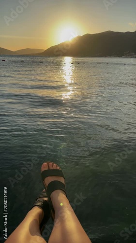 Female legs above calm sea at sunset in Montenegro, with golden light, mountain silhouettes and peaceful evening atmosphere by the coast