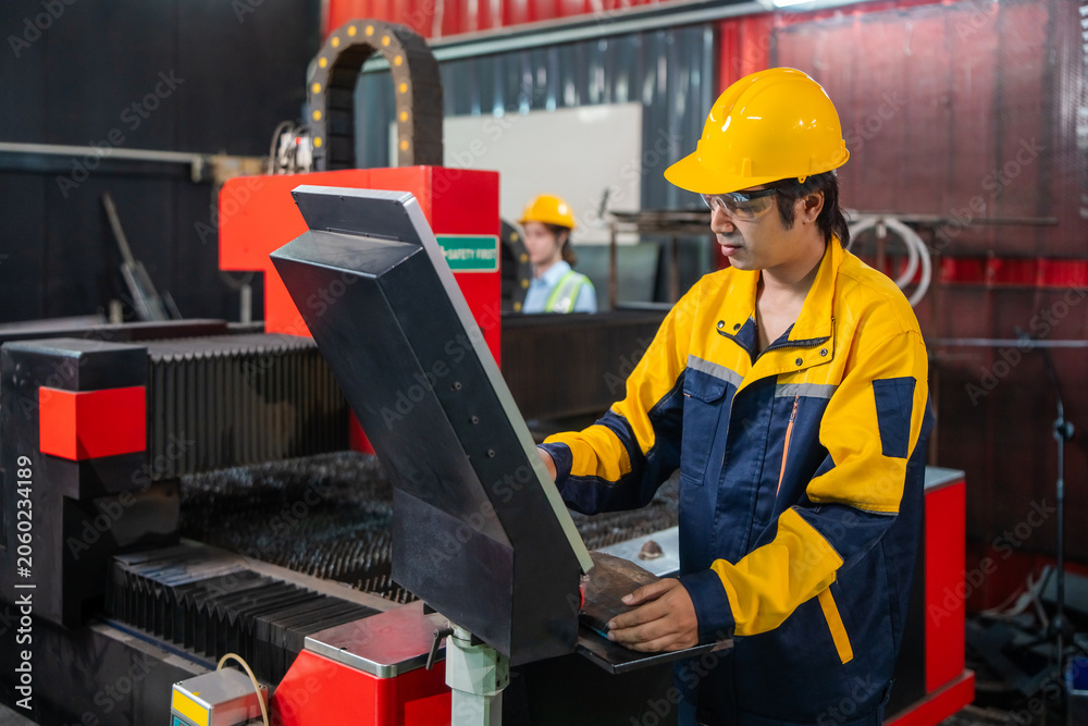 Fototapeta premium Worker in a yellow and blue work jacket operates a large industrial machine. They wear a yellow hard hat and use a control panel. Another worker in a yellow hard hat is visible in the background