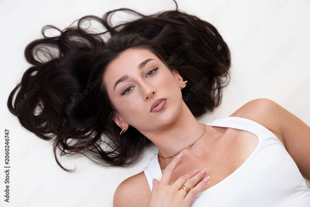 Naklejka premium Beauty and fashion portrait of a young woman with long dark hair, natural makeup, and elegant jewelry. The female model is lying down, looking directly at the camera, her features and accessories. Thi