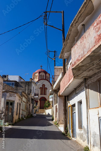 Picturesque narrow street in Gouves village, Crete, Greece, with the historic Zoodochos Pigi church visible in the background under a sunny sky