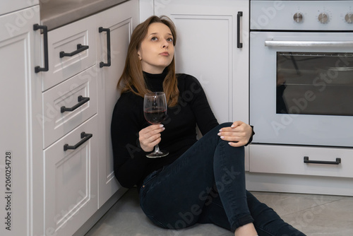 Bild auf Leinwand Young woman sitting on kitchen floor, sipping wine, struggling with feelings of