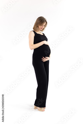 Happy pregnant woman in a black outfit gently holding her belly on white background. Vertical photo.