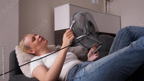 Woman lying on sofa in home during extreme heat wave, cooling her face with electric fan and feeling relief from dangerously hot indoor temperature.extreme heat,  climate heatwave concept