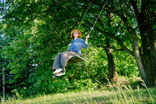 Happy woman in a hat and sunglasses swinging on a swing in a sunny summer forest