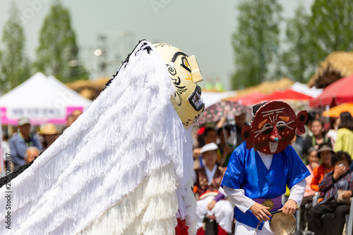 Korean traditional mask dance performance at an outdoor festival with colorful costumes and audience in the background.