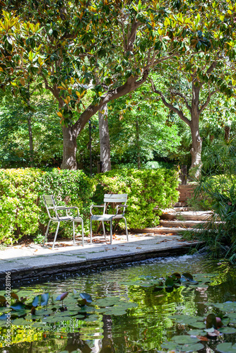 A tranquil garden scene featuring a small pond with blooming water lilies, comfortable chairs beside the water, lush green hedges, and mature magnolia trees creating natural shade. 