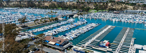 The California, Dana Point Harbor,  with Bots and Yachts in their slips as people interact with the pacific ocean and sea cliffs in the background and sailboats