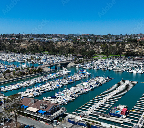 The California, Dana Point Harbor,  with Bots and Yachts in their slips as people interact with the pacific ocean and sea cliffs in the background and sailboats