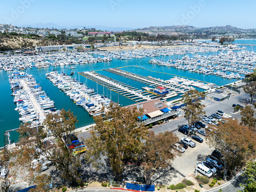 The California, Dana Point Harbor,  with Bots and Yachts in their slips as people interact with the pacific ocean and sea cliffs in the background and sailboats