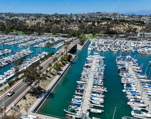 The California, Dana Point Harbor,  with Bots and Yachts in their slips as people interact with the pacific ocean and sea cliffs in the background and sailboats