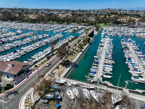 The California, Dana Point Harbor,  with Bots and Yachts in their slips as people interact with the pacific ocean and sea cliffs in the background and sailboats