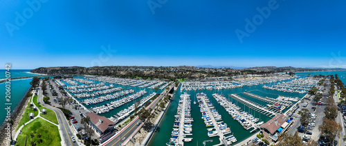 The California, Dana Point Harbor,  with Bots and Yachts in their slips as people interact with the pacific ocean and sea cliffs in the background and sailboats