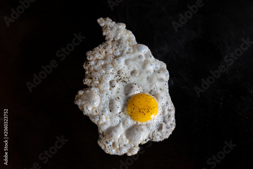 sunny side up egg with sprinkle of black paper and salt cooking on a hot flat top griddle in a commercial kitchen