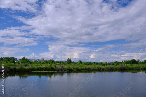 Calm water surface lake reflecting bright clouds with green trees and buildings far away.