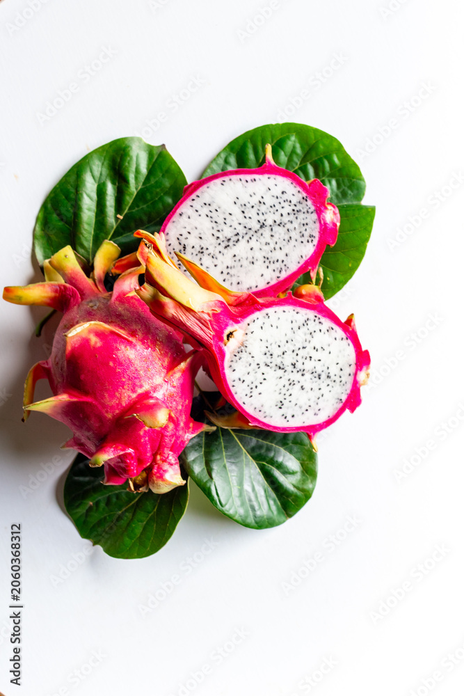 Fototapeta premium Two pitaya fruits - whole and cut in half - with green leaves on white table. Ripe dragon fruit or pitahaya. Tropical fruits, top view