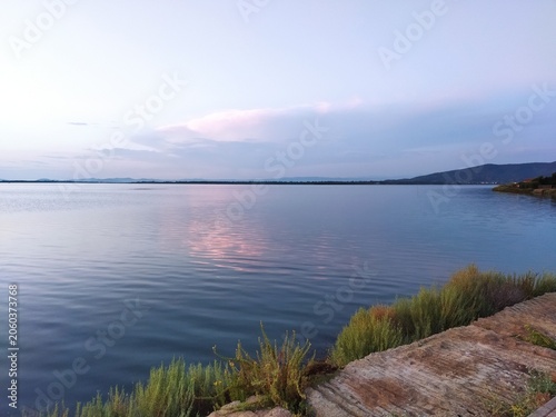 Atardecer en la Laguna de Orbetello con reflejos rosados en el agua tranquila y vegetación en la orilla, Toscana, Italia.