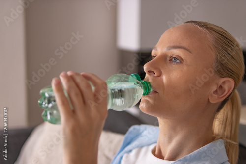 Tableau sur toile Woman sitting on sofa at home drinking water from bottle, relaxing and rehydrating in comfortable living room after a warm day, enjoying calm indoor lifestyle moment