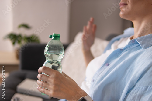 фотографія Woman sitting on sofa at home drinking water from bottle, relaxing and rehydrating in comfortable living room after a warm day, enjoying calm indoor lifestyle moment