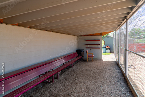 Foto Empty baseball dugout interior with weathered red benches, cinder block wall, and gravel floor, looking out onto a sports field through a chain-link fence on a sunny day