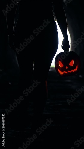 Vertical video. Silhouette Walking Through Dark Tunnel Carrying Glowing Jack O Lantern Toward Bright Light at Tunnel End, Halloween Mystery, Fear And Unknown Journey