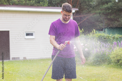Young man in glasses spraying water from a garden hose with a nozzle on a lawn, dynamic water mist.