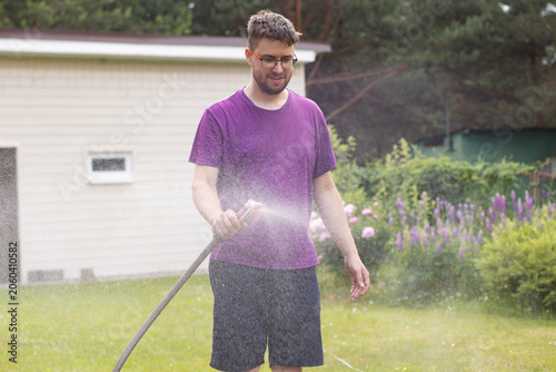 Young man in glasses spraying water from a garden hose with a nozzle on a lawn, dynamic water mist.