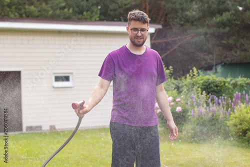 Young man in glasses spraying water from a garden hose with a nozzle on a lawn, dynamic water mist.