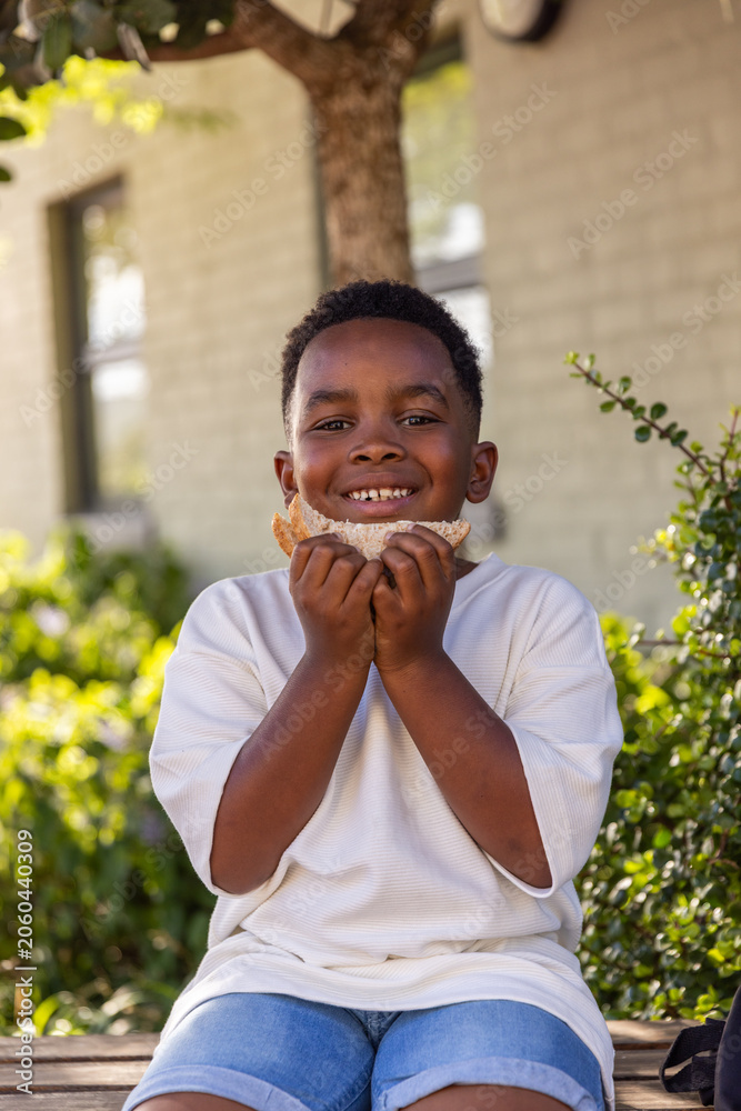 Fototapeta premium African American child sitting on bench in front yard holding sandwich in white tee, denim shorts