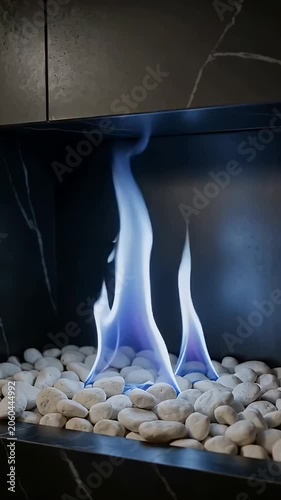 Blue flames rise from white pebbles in a modern fireplace with dark marble surround and sleek black interior.