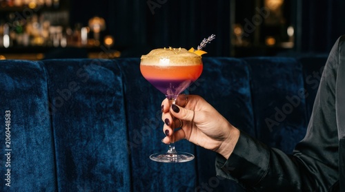Woman's hand holding a layered purple and orange cocktail with gold glitter garnish in a dimly lit upscale bar setting
