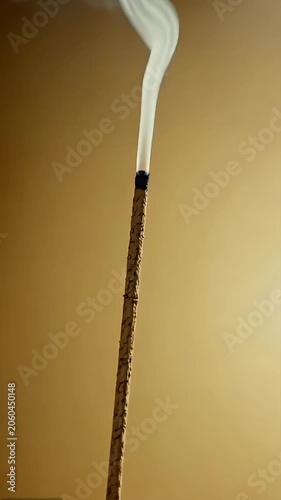 Single burning incense stick releasing white smoke against a plain beige wall background in vertical composition.
