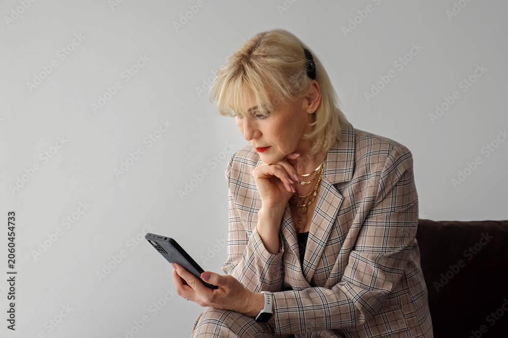 Naklejka premium Senior woman on a couch focused on her smartphone, quietly engaging with digital content and messaging at home in a smart casual plaid blazer and accessories