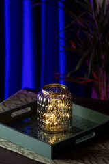 "Glass jar with warm fairy lights on decorative tray and blue background"