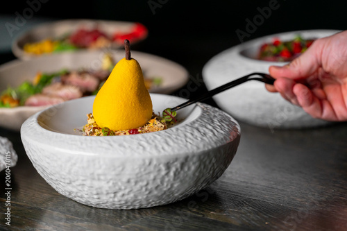 Tableau sur toile A bright yellow pear-shaped dessert sits in a white textured bowl on a dark table, as a hand places a garnish next to it using tweezers