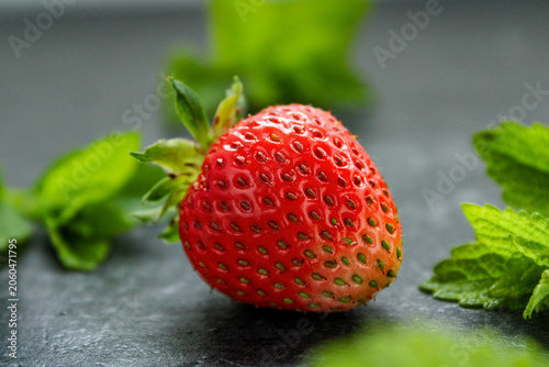 Single ripe strawberry with fresh mint leaves on a dark slate background, featuring vibrant red color and selective focus macro photography.