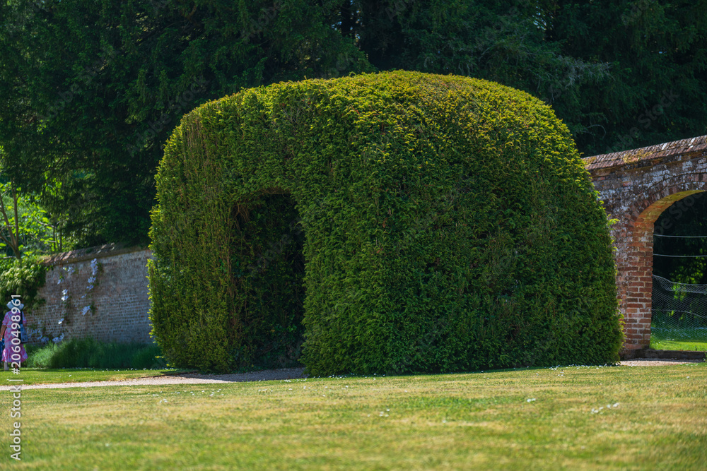 Fototapeta premium Highclere, United Kingdom - 21.05.2026: Large clipped topiary arch beside brick garden arcade on estate grounds, with a distant visitor visible.