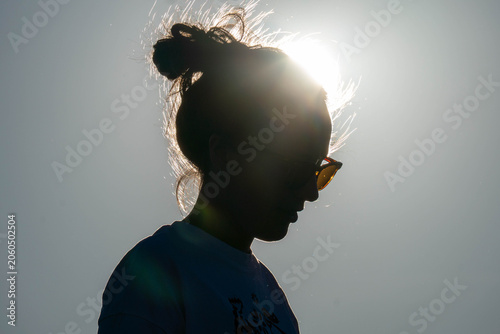 Silhouette of a woman in sunglasses with a messy bun, backlit by bright sun against a clear sky, creating a dramatic summer profile.