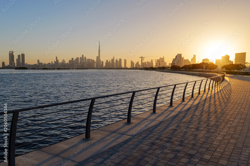 Obraz premium Sunset view from Al Jaddaf Walk waterfront promenade overlooking Dubai skyline and Burj Khalifa, with golden evening light reflecting across Dubai Creek