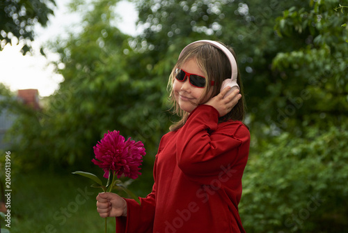 Slika na platnu Portrait of a happy girl in headphones with a burgundy peony in her hand against a picturesque green garden background